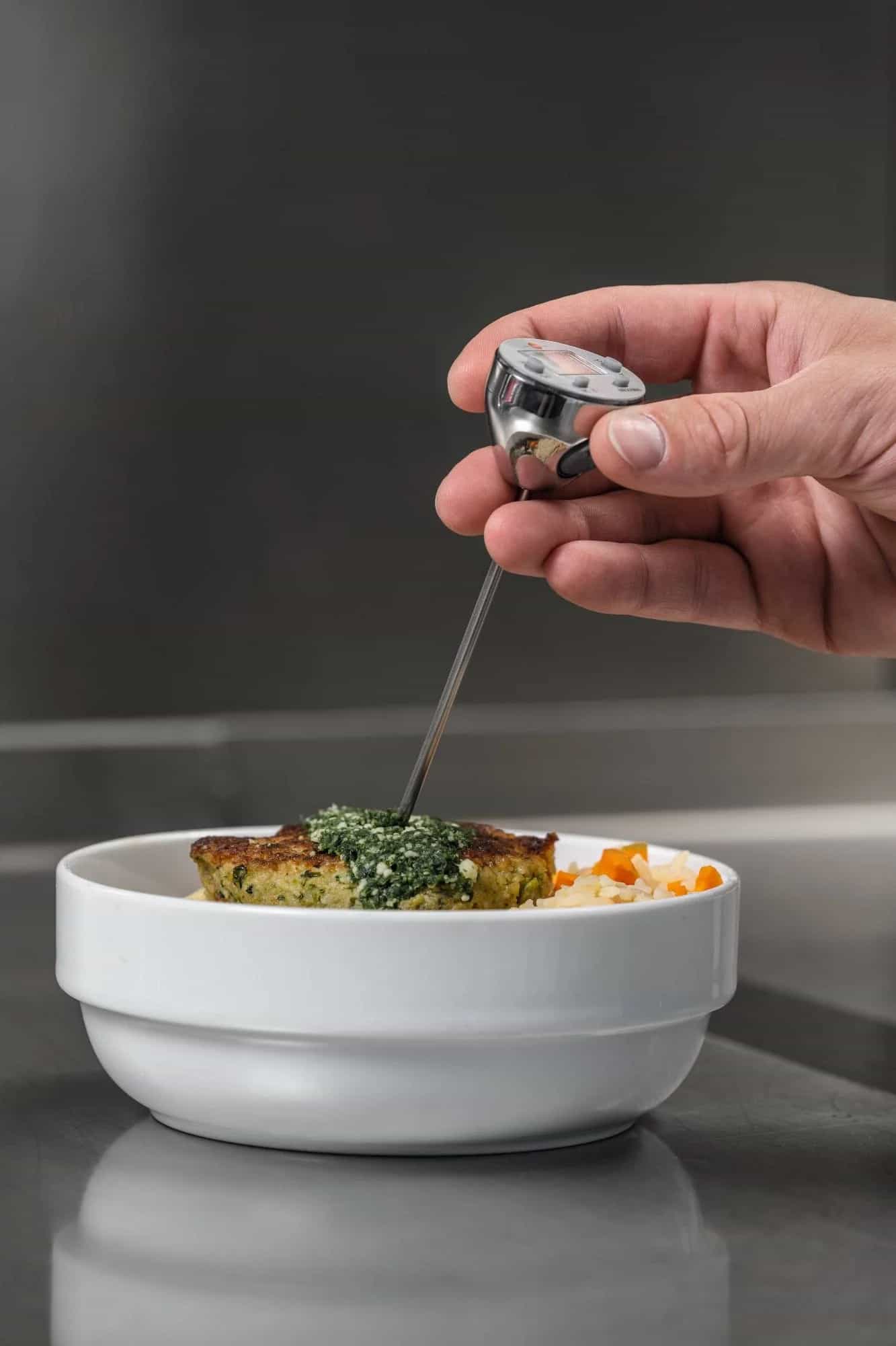 Dłoń wbija sondę minitermometru Testo w danie mięsno-warzywne na talerzu — kontrola temperatury gotowego posiłku HACCP (miniatura)