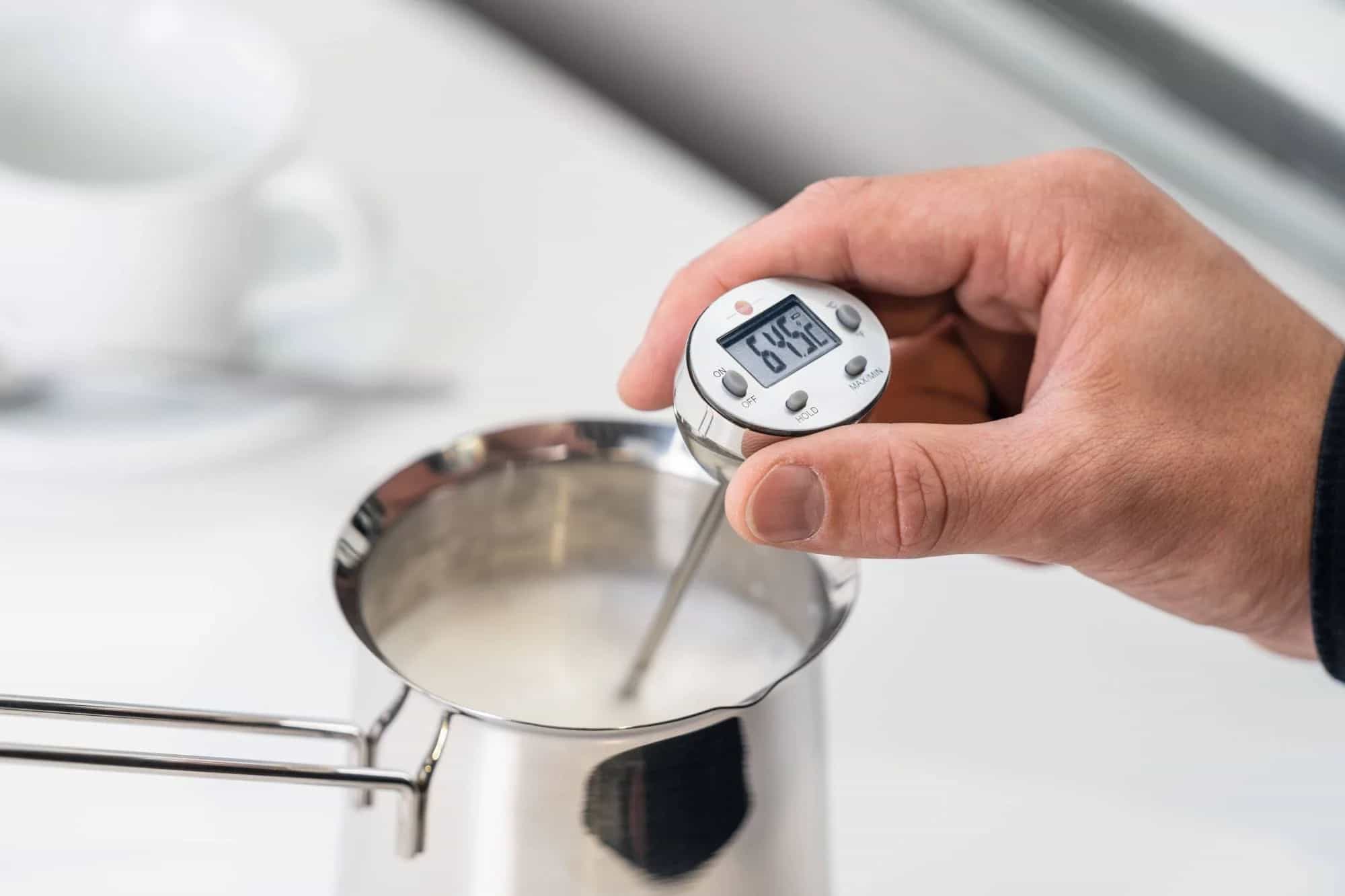 Dłoń trzyma minitermometr Testo zanurzony w mleku w garnku ze stali nierdzewnej — pomiar temperatury w gastronomii (miniatura)