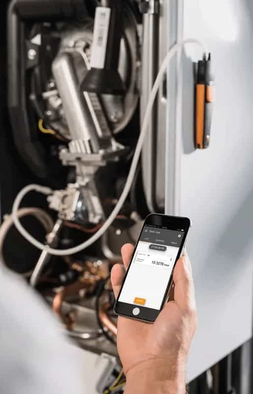 Testo 510i podczas pomiaru na instalacji grzewczej, technik trzyma smartfon z uruchomioną aplikacją Testo Smart App (miniatura)