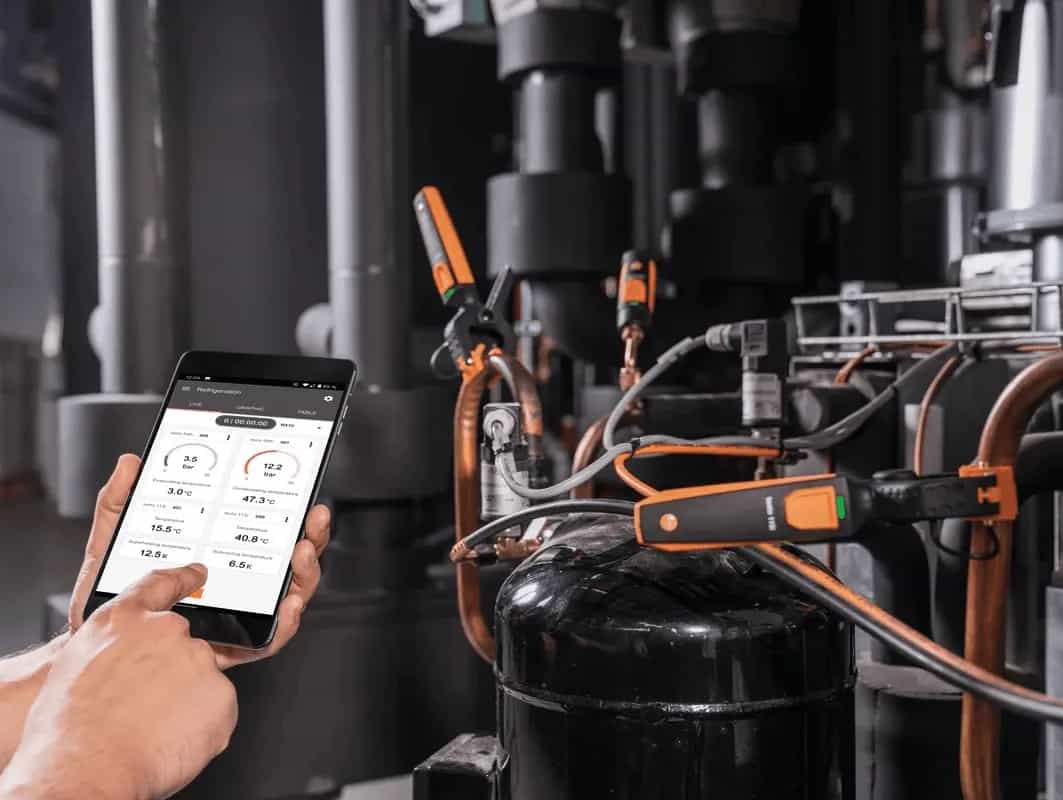 Termometr zaciskowy Testo 115i zamontowany na rurach instalacji chłodniczej, z widoczną aplikacją Testo Smart App na smartfonie (miniatura)