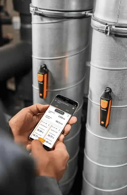 Dłonie trzymające smartfon z aplikacją Testo Smart App przy metalowych kanałach wentylacyjnych (miniatura)