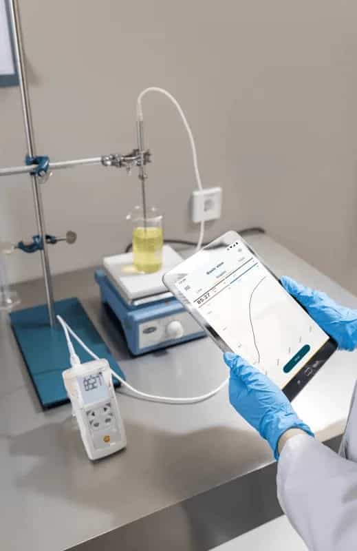 Badacz w rękawiczkach laboratoryjnych obsługuje tablet z wykresem temperatury przy stanowisku z mieszadłem magnetycznym i miernikiem Testo 110 (miniatura)