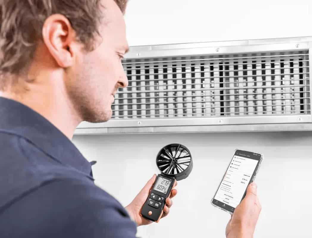 Technik odczytujący pomiar z anemometru Testo 417 oraz z aplikacji Testo Smart App na smartfonie przy kratce wentylacyjnej (miniatura)