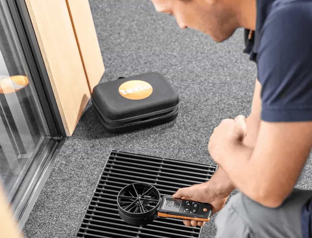 Serwisant mierzący przepływ powietrza anemometrem Testo 417 przy podłogowej kratce wentylacyjnej (miniatura)