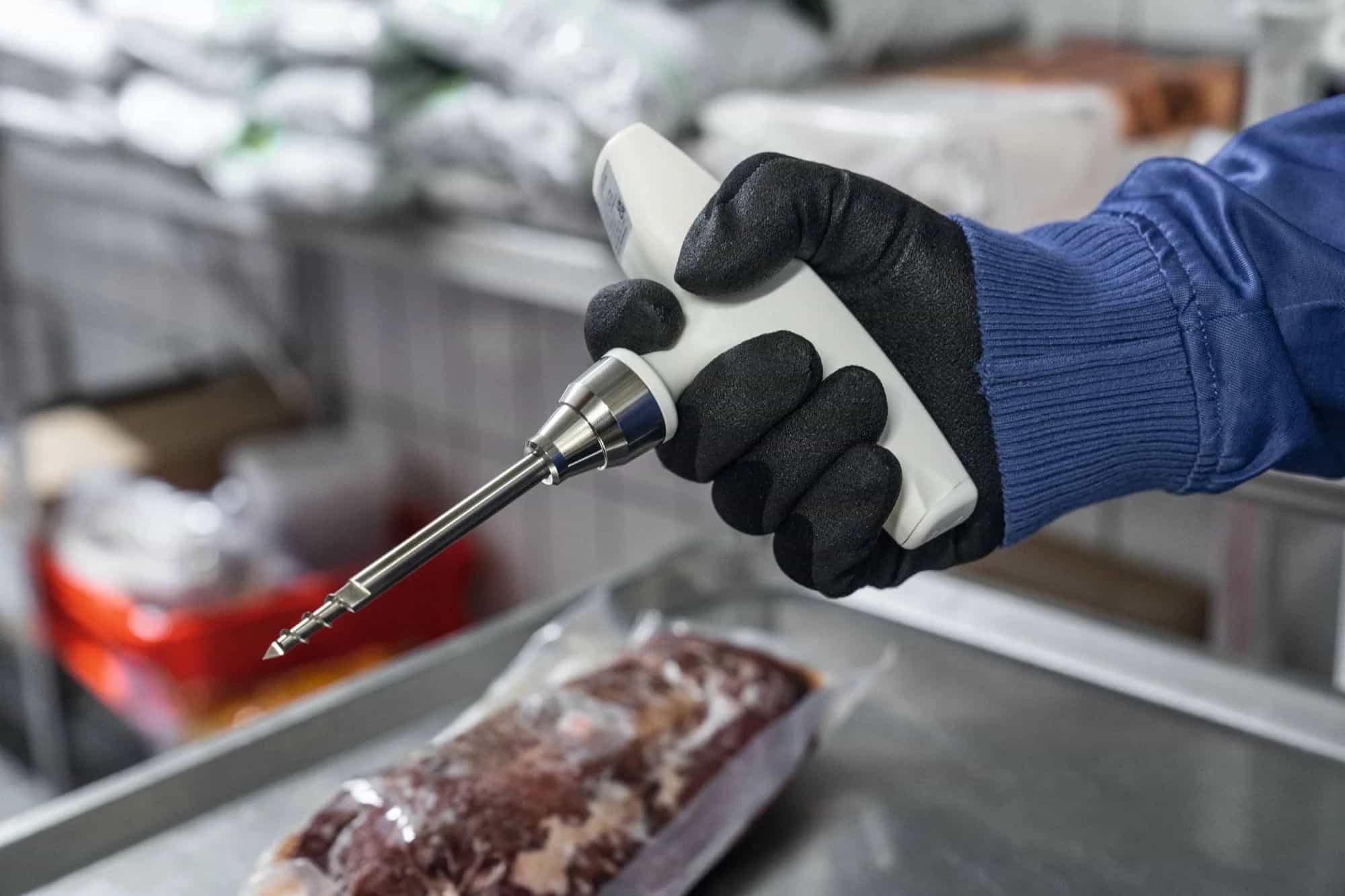 Pracownik w rękawicy trzymający termometr Testo 105 podczas pomiaru temperatury mrożonego mięsa w magazynie (miniatura)