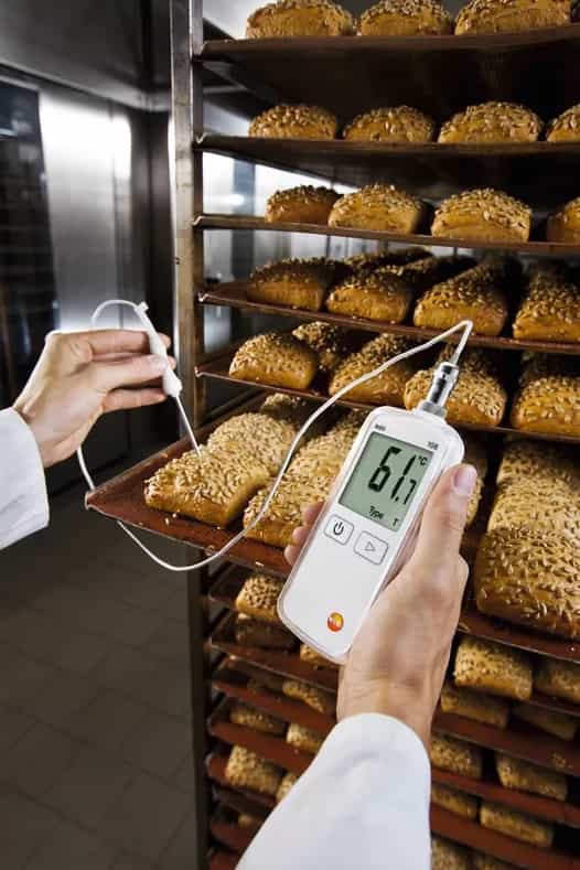 Testo 108-2 podczas pomiaru temperatury bułek w piekarni — dłonie operatora i regał z pieczywem (miniatura)