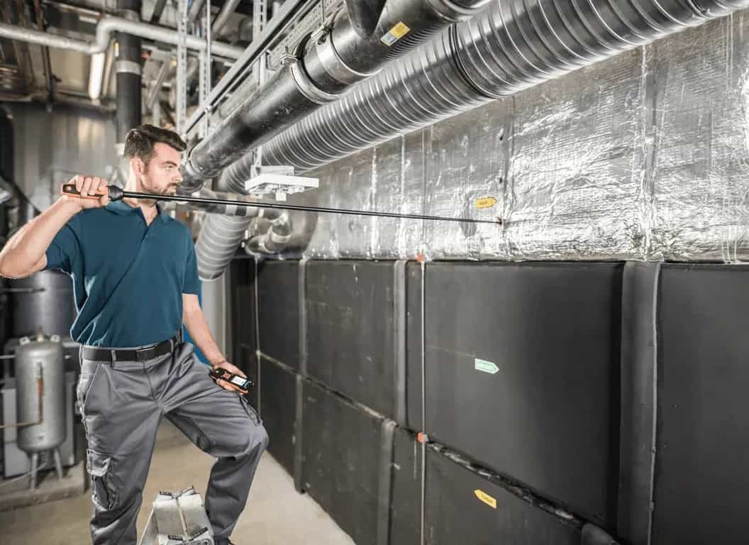 Technik wykonujący pomiar przepływu i temperatury powietrza miernikiem Testo 440 przy instalacji wentylacyjnej w hali technicznej (miniatura)