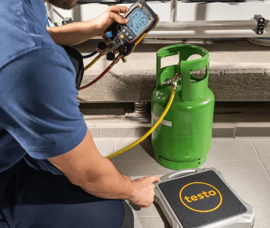 Technik mierzy masę czynnika chłodniczego wagą Testo 560i przy zielonej butli, obsługując pomiar na tablecie (miniatura)