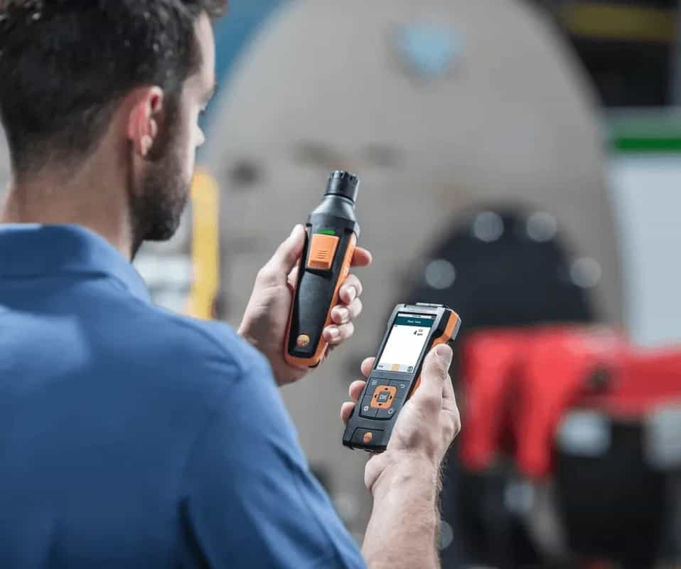 Technik trzymający cyfrową sondę Testo do pomiaru CO obok smartfona z aplikacją Testo na tle kotła (miniatura)