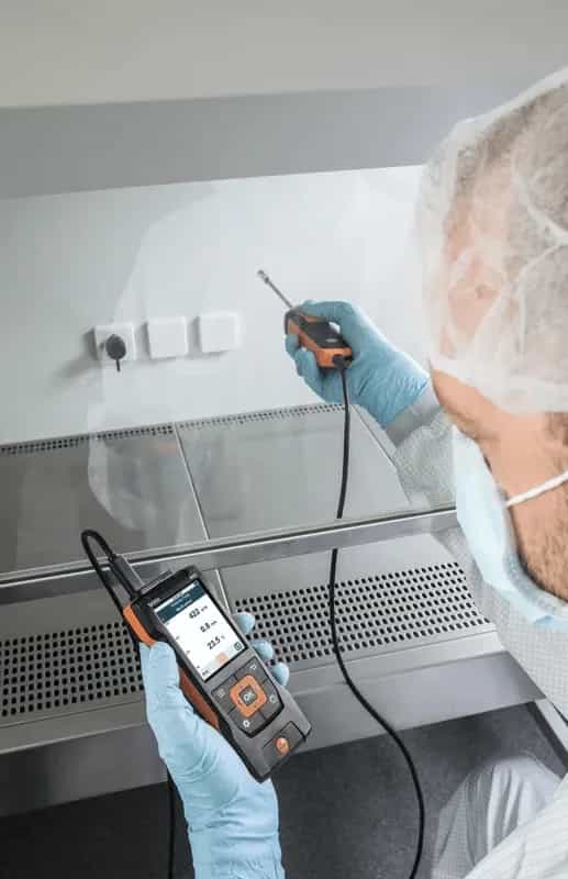 Technik laboratoryjny w fartuchu i czepku wykonujący pomiar przy szybie dygestorium sondą Testo (miniatura)