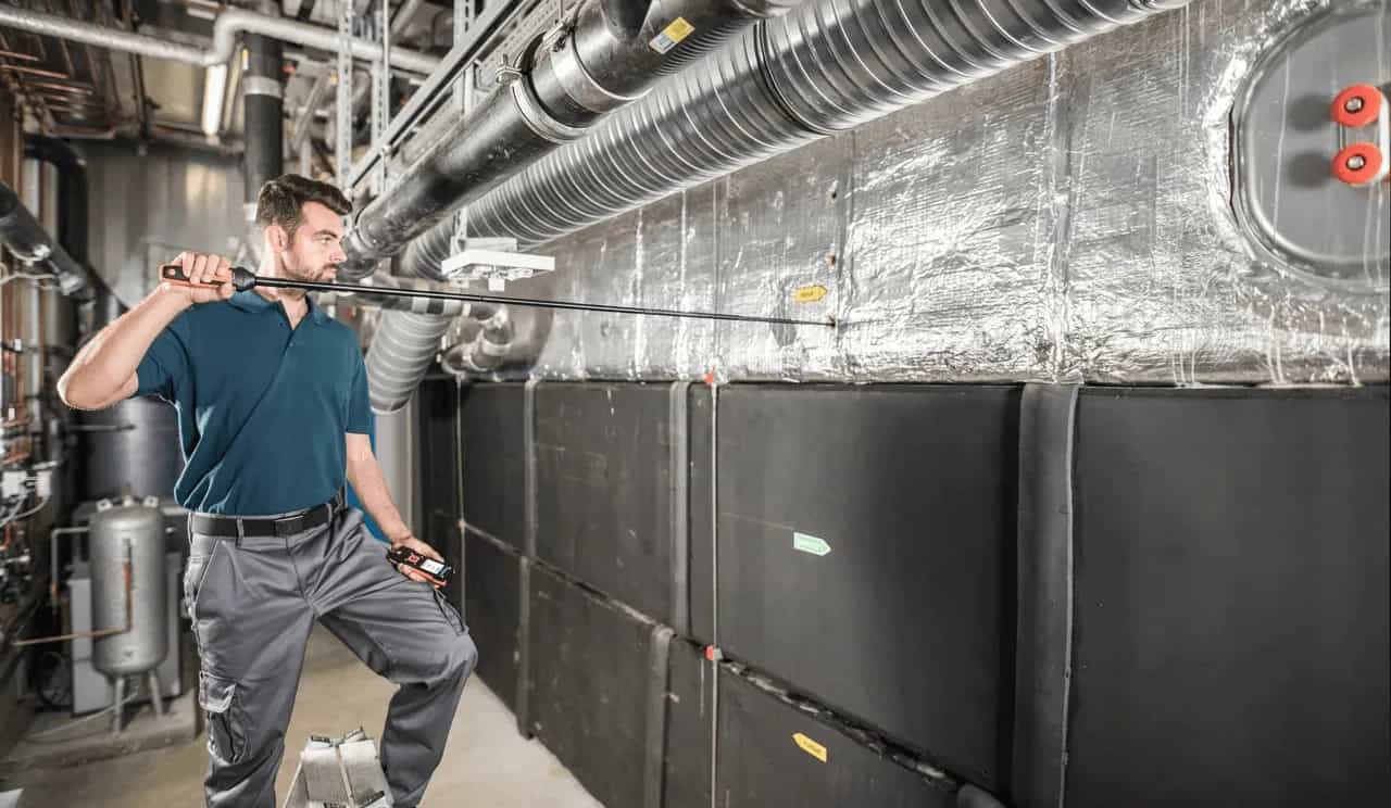 Technik HVAC przeprowadzający pomiar sondą Testo przy dużym agregacie wentylacyjnym w hali przemysłowej (miniatura)