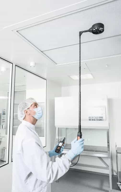 Technik w odzieży ochronnej mierzący przepływ powietrza przy suficie pomieszczenia czystego za pomocą sondy wiatraczkowej Testo Ø 100 mm na teleskopie (miniatura)