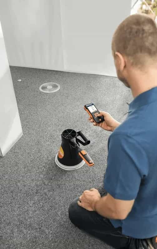 Technik mierzy przepływ powietrza w kanale wentylacyjnym przy suficie, trzymając sondę wiatraczkową Testo na wysięgniku i miernik Testo w drugiej ręce (miniatura)