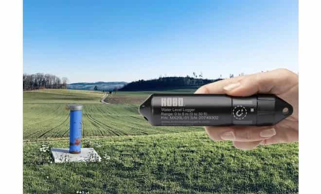 HOBO MX20L-02 trzymany w dłoni na tle pola z piezometrem — montaż w studni (miniatura)