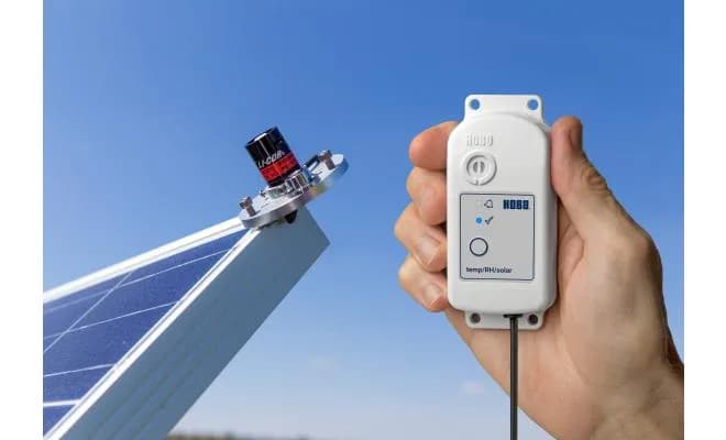 Rejestrator HOBO MX2309 zamontowany przy instalacji fotowoltaicznej — czujnik LI-COR LI-200R na panelu słonecznym, użytkownik trzyma rejestrator na tle nieba