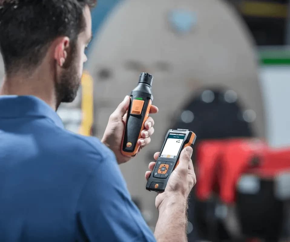 Technik trzymający sondę Testo do pomiaru CO i smartfon z aplikacją połączoną przez Bluetooth w pomieszczeniu technicznym (miniatura)