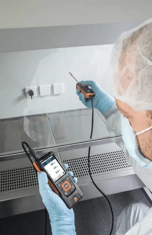 Technik laboratoryjny w fartuchu i czepku wykonujący pomiar przy szybie dygestorium sondą Testo (miniatura)