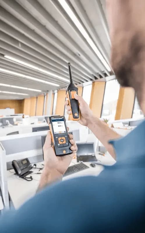 Technik odczytuje pomiar z miernika Testo i aplikacji mobilnej połączonej przez Bluetooth podczas kontroli klimatu w pomieszczeniu biurowym (miniatura)