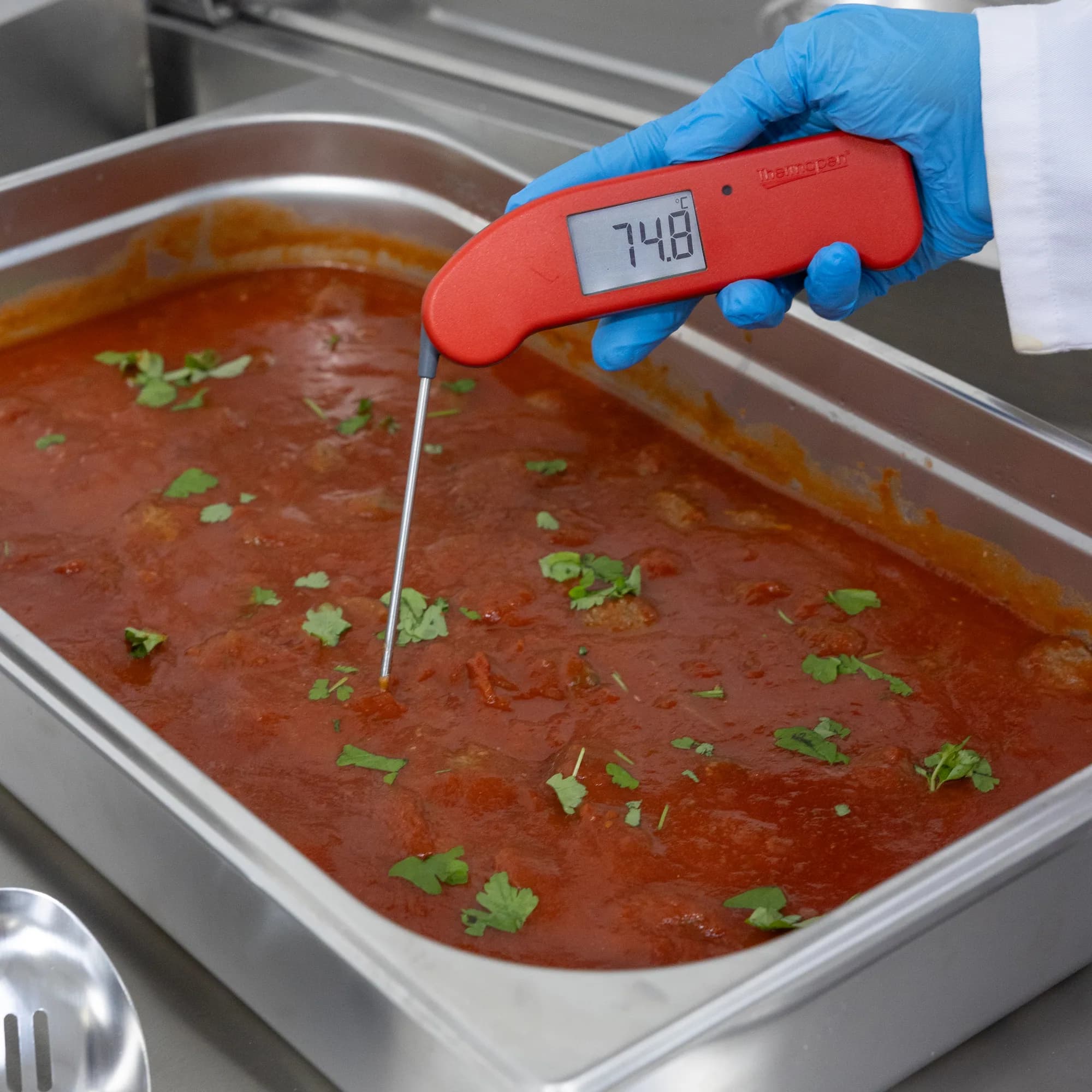 Kucharz w rękawiczce mierzy termometrem szpilkowym temperaturę potrawy w pojemniku gastronomicznym, odczyt 74,8 °C