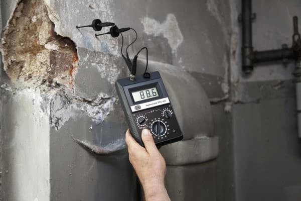 Miernik Gann RTU 600 w dłoni podczas pomiaru w uszkodzonym tynku, z elektrodami osadzonymi w murze
